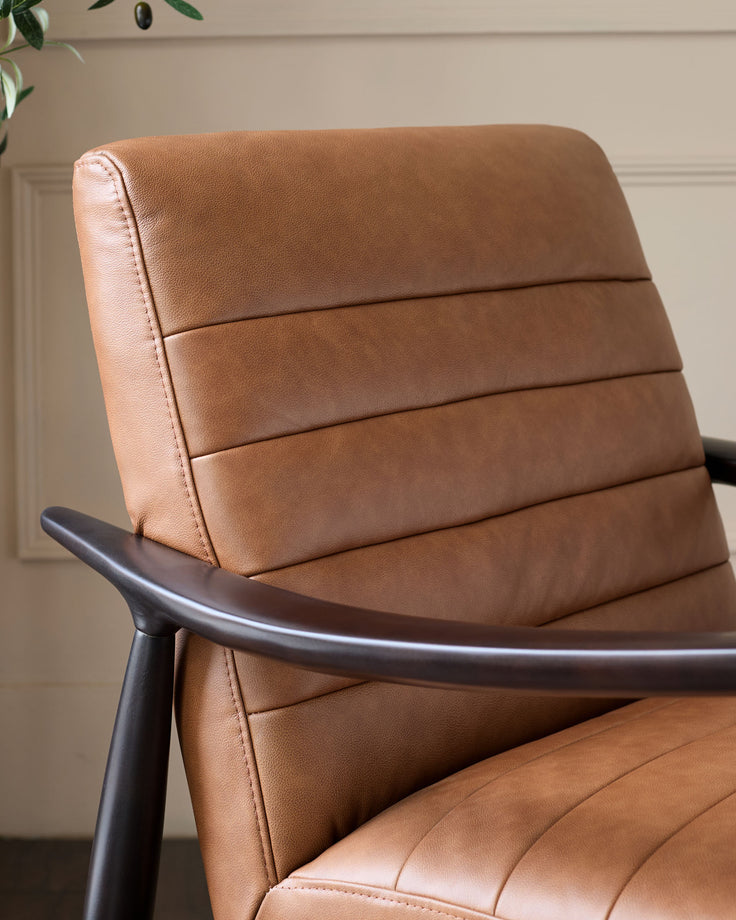 Tan leather accent chair with horizontal channel stitching and curved dark metal armrest, photographed at an angle.
