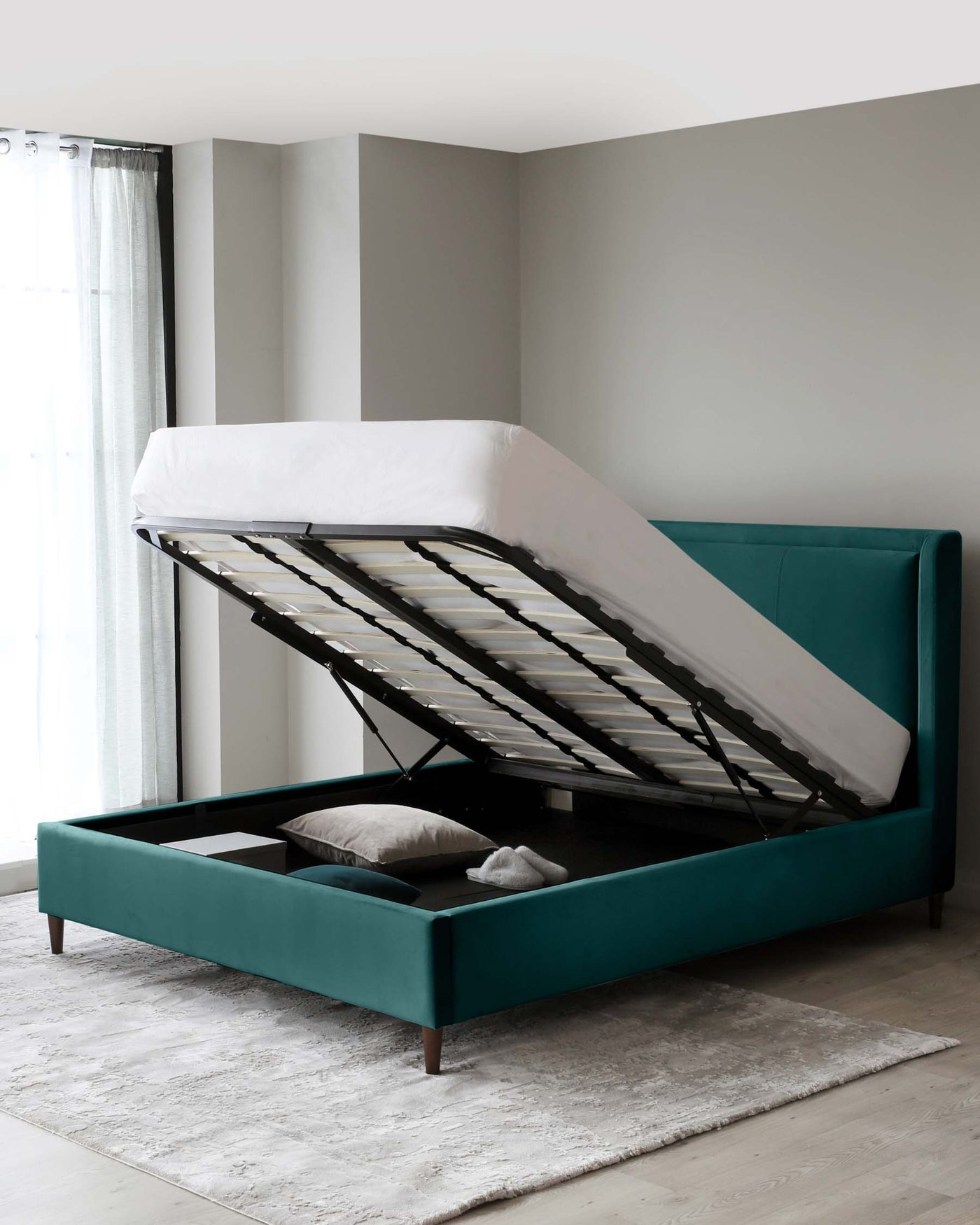 A modern teal upholstered storage bed with a lifted mattress base revealing a spacious storage compartment underneath, set against a neutral backdrop with a soft grey area rug on the floor.