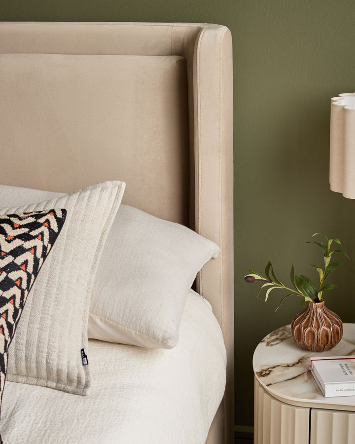 Champagne velvet bed headboard with cream bedding, patterned pillow, and marble side table with small vase against green wall.