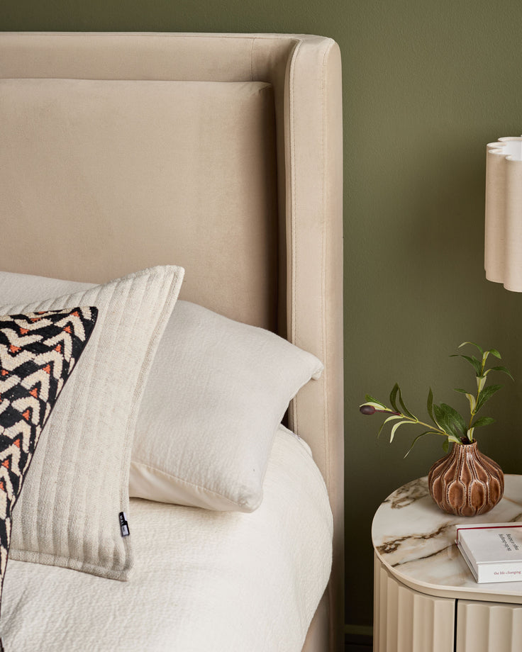 Champagne velvet bed headboard with cream bedding, patterned pillow, and marble side table with small vase against green wall.