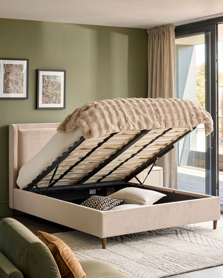 Grove King Ottoman Bed in champagne velvet with lifted storage base, displayed in a bedroom with sage green walls.