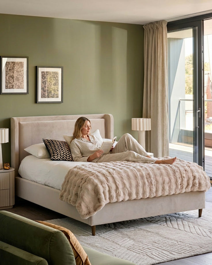 Grove King Ottoman Bed in champagne velvet, displayed in a bedroom setting with a woman reading on the bed.