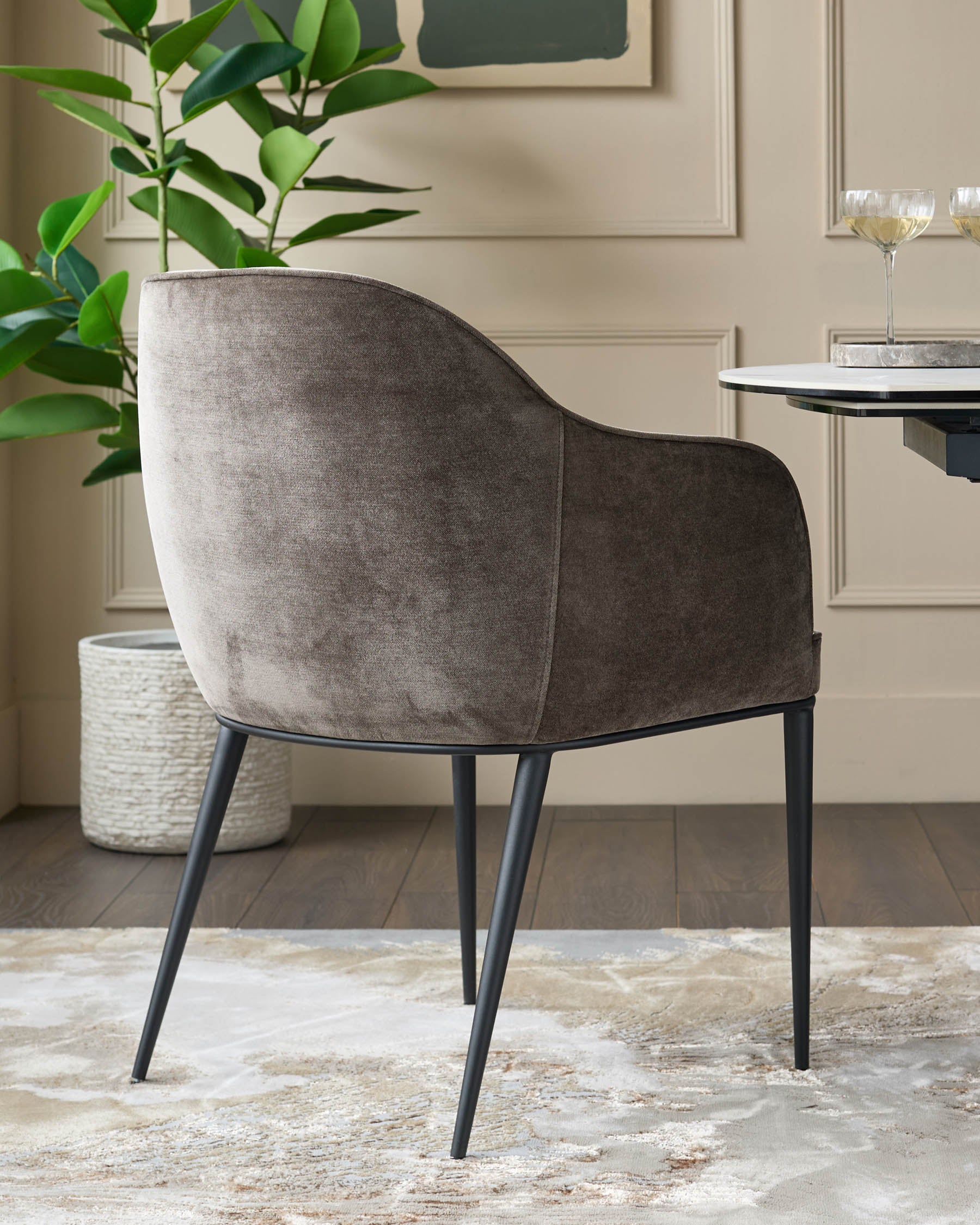 Upholstered dining chair in brown velvet with black legs beside a round marble table with wine glasses.