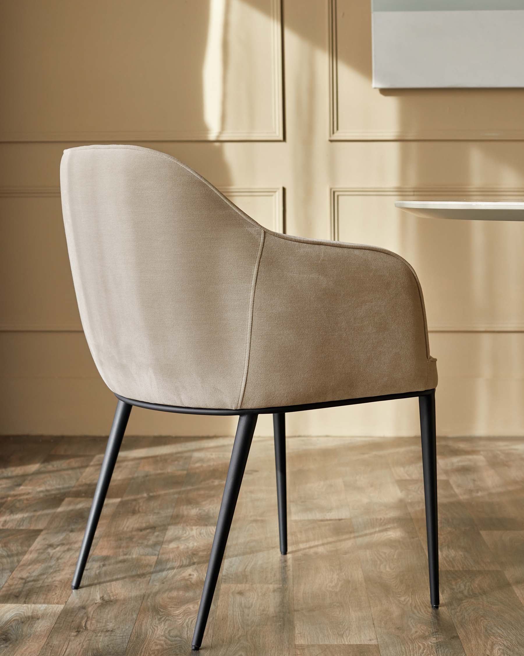 A modern beige chair with black metal legs sits on a wooden floor against a beige panelled wall.