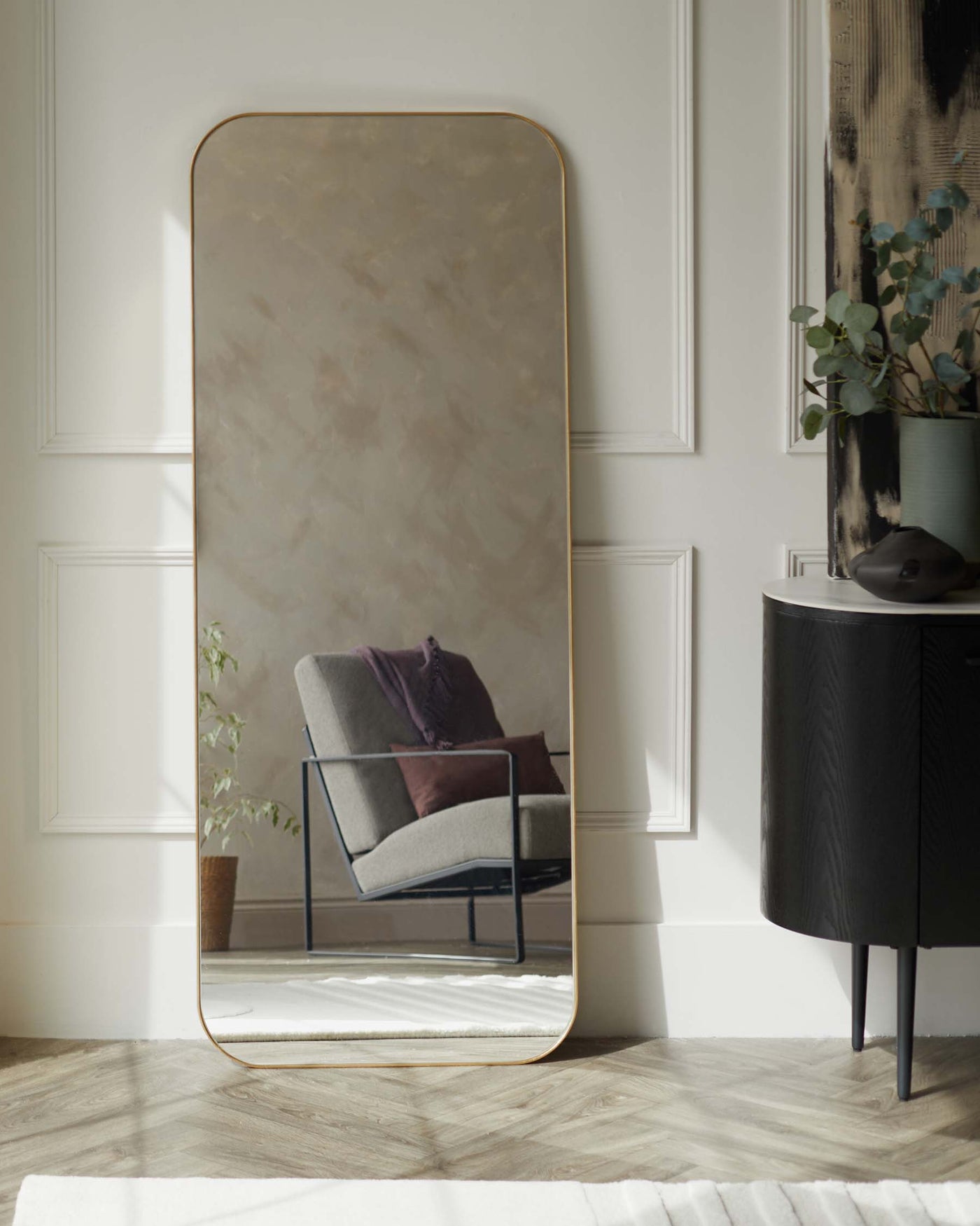 Large floor-standing mirror with a rounded top and gold-finished metal frame, and a sleek black round side table with ribbed texture and thin metal legs, reflected in the mirror.