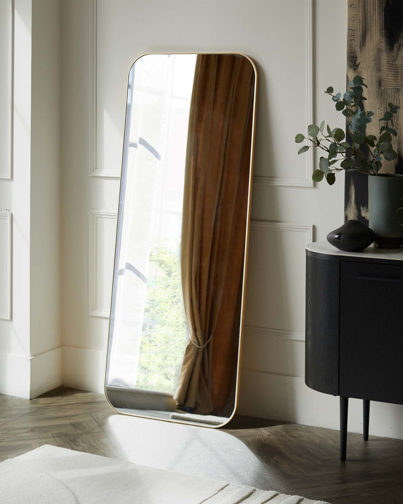 Large full-length floor mirror with a rounded top and brass-finished frame, alongside a dark-toned wooden sideboard with round tapered legs and sophisticated textured surface detailing.