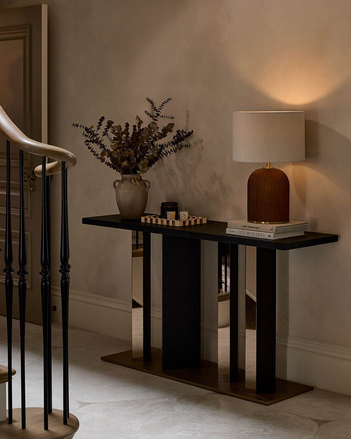 Black console table with geometric base design, styled with decorative items including a vase, lamp, and books in a modern entryway.