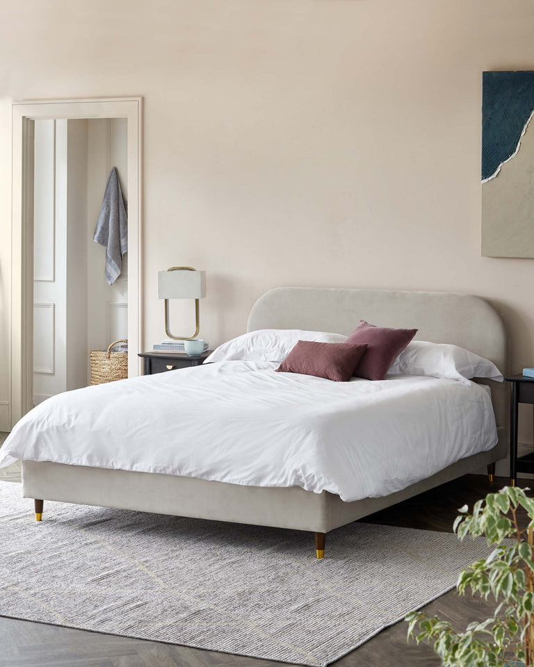 Elegant modern bedroom featuring a plush upholstered bed with a curved headboard and wooden legs with brass caps, accompanied by a sleek midnight blue nightstand with an artistic table lamp. A woven grey area rug sits under the bed, adding warmth to the space.