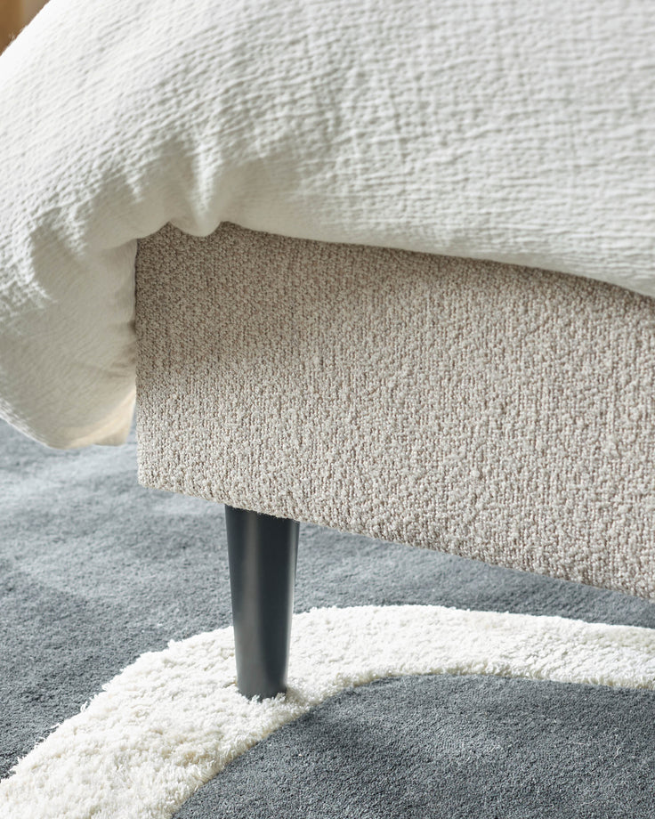 Close-up of a sofa corner with textured upholstery, supported by a dark leg on a patterned rug.