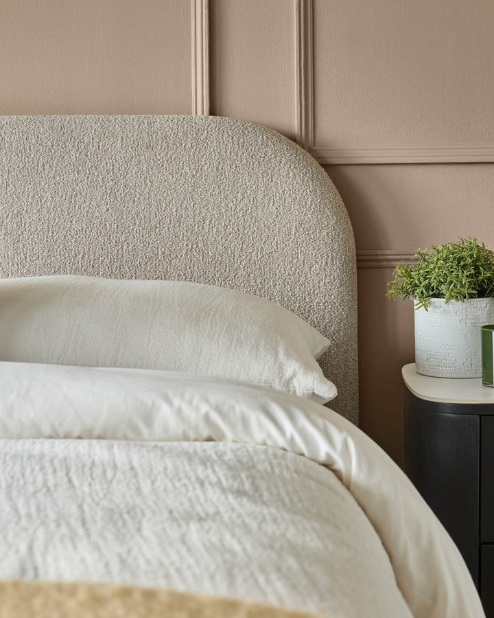 Bedroom featuring a textured headboard, cream bedding, dark bedside table and decorative plant.