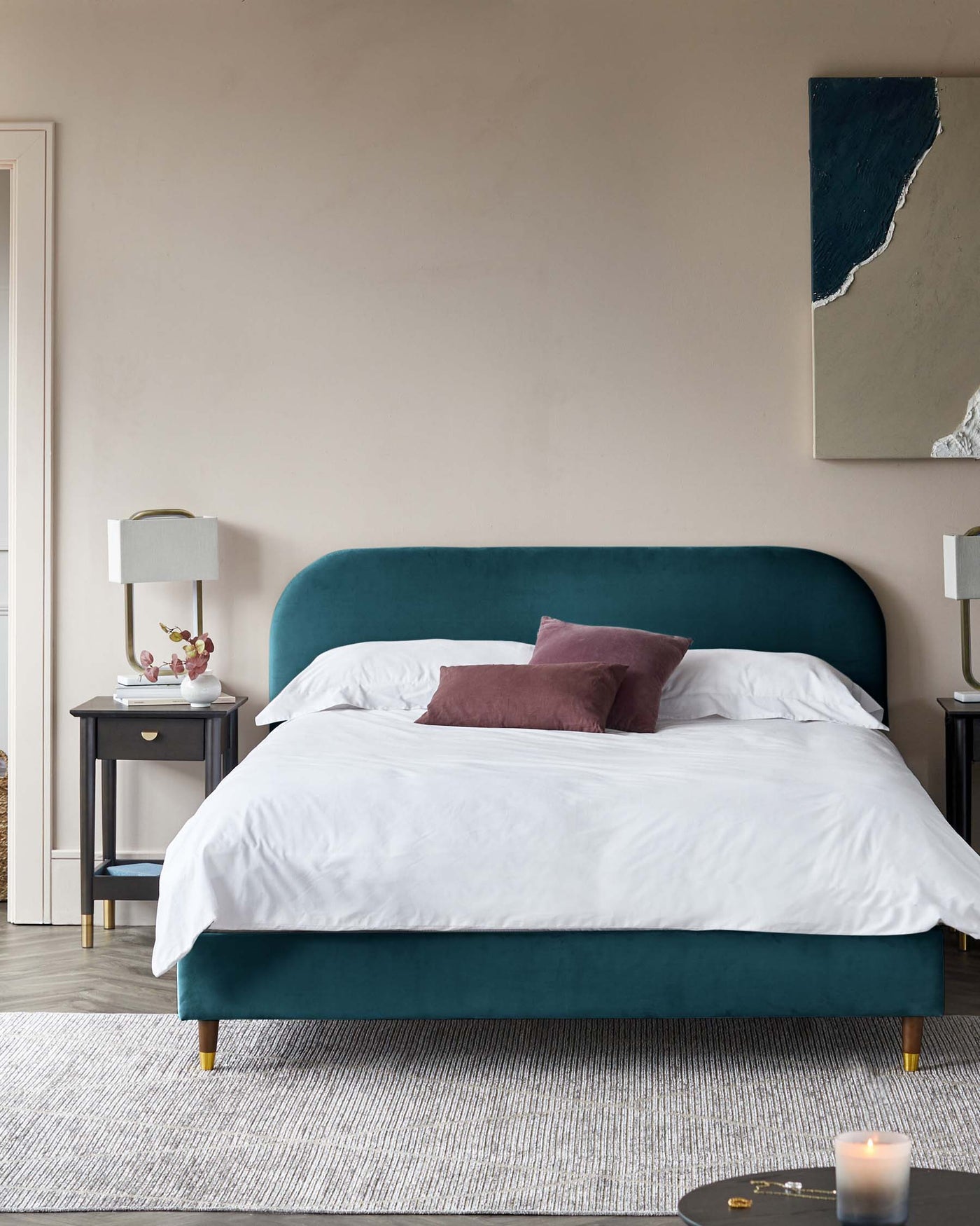 A luxurious teal upholstered bed with a curved headboard and brass-capped legs, flanked by two navy blue nightstands with brass knobs and a lower shelf, set on a textured off-white area rug. A white lamp with a cylindrical shade rests on one nightstand, and an abstract painting hangs above the bed.