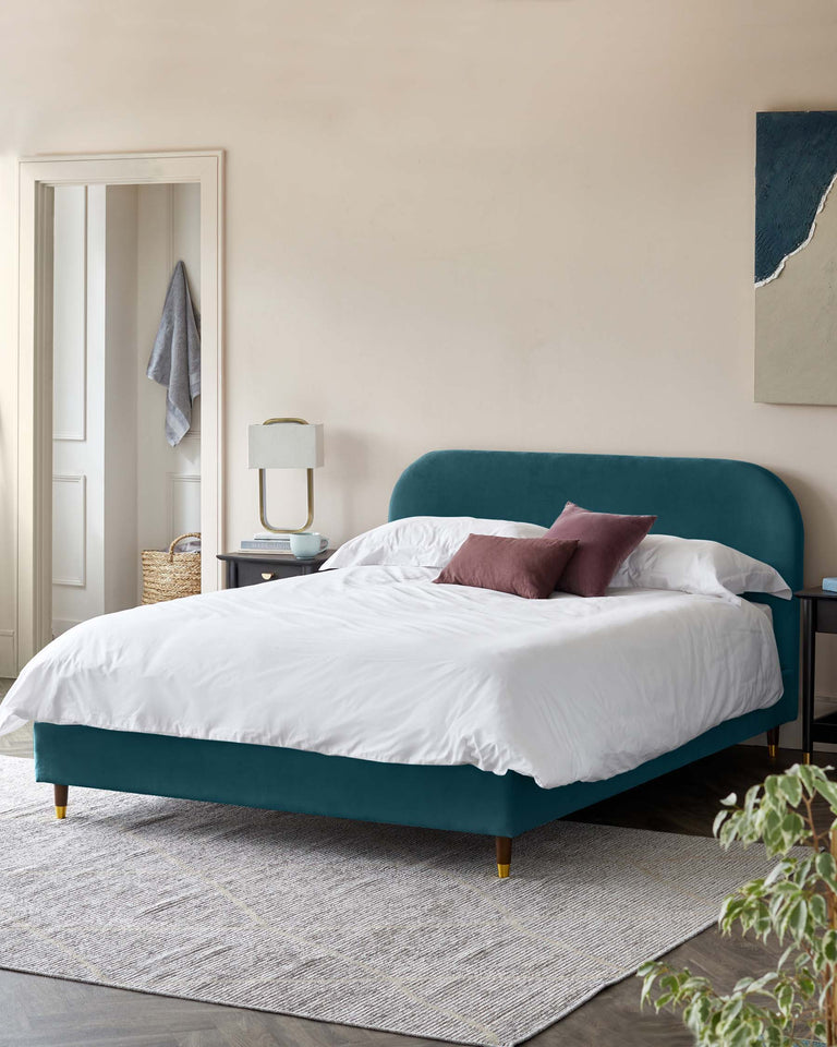 Elegant contemporary bedroom featuring a teal upholstered platform bed with a curved headboard and contrasting wooden legs tipped with gold accents. Bed is dressed in white bedding with contrasting plum accent pillows. A minimalist white and blue bedside table with mid-century modern design elements holds a ceramic table lamp. The arrangement is completed with a soft textured area rug in neutral tones.