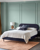 A bedroom with a plush, dark blue bed, white linen, side tables, and a patterned rug. A calming and stylish space.