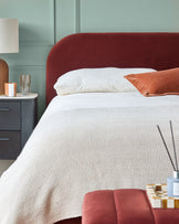 Bedroom scene: upholstered bed with a bedside table, lamp, candle, & ottoman. Cosy textiles add warmth.