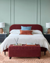 Bedroom with a deep red bed and ottoman, bedside tables, lamps, and a patterned rug, creating a cosy, stylish interior.