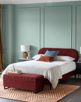 A bedroom with a red bed, ottoman and rug. There are two bedside tables and lamps on either side of the bed.