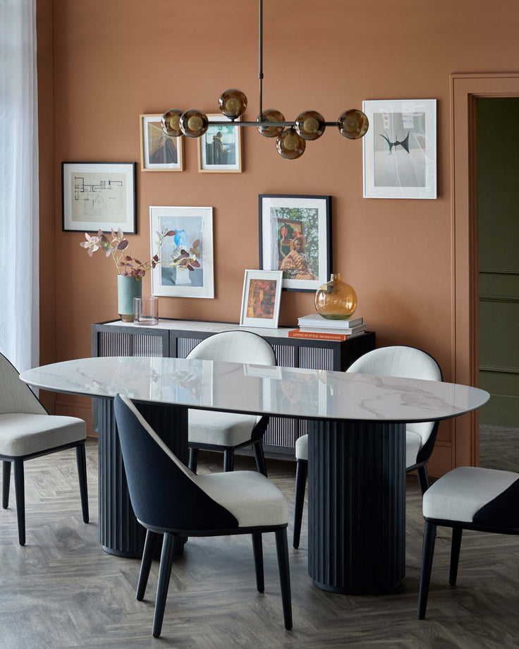 Modern dining table with oval marble top, black fluted base, and upholstered chairs in two-tone fabric. Gallery wall and pendant light.
