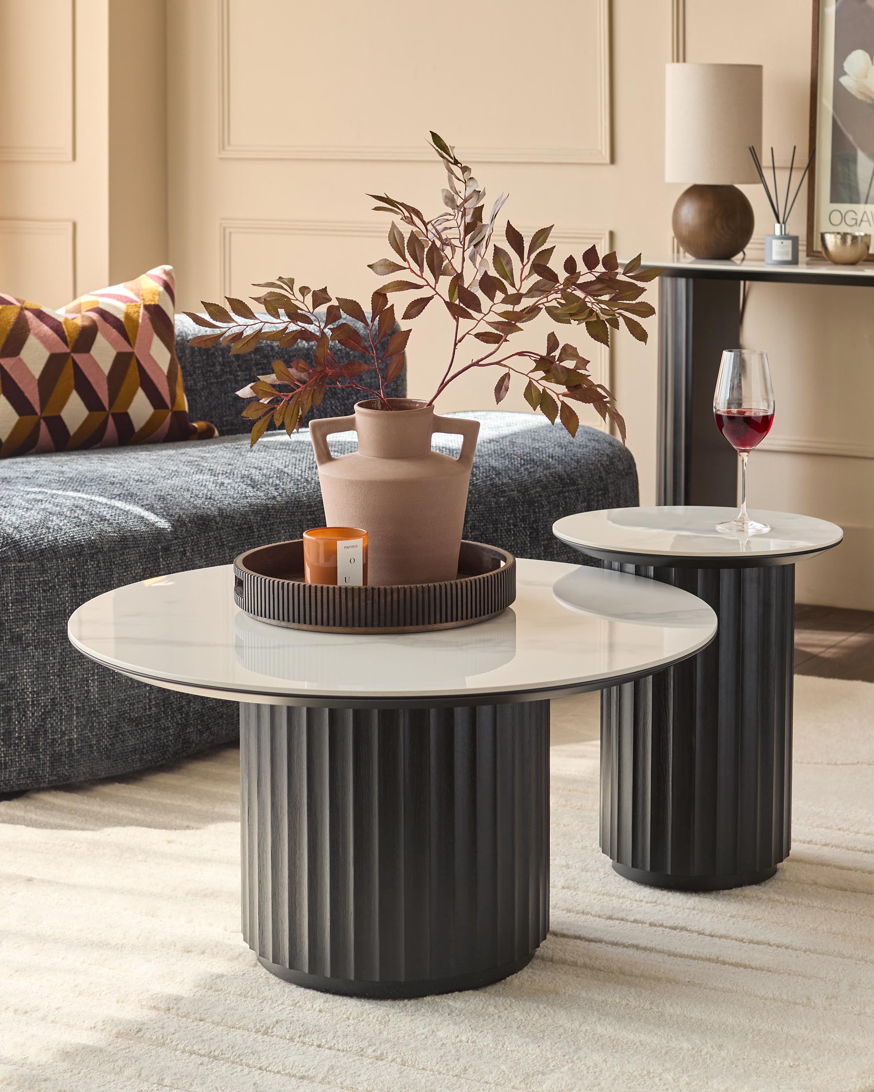 Two marble-topped coffee tables with dark, fluted bases sit on a cream rug next to a grey sofa and other decor.