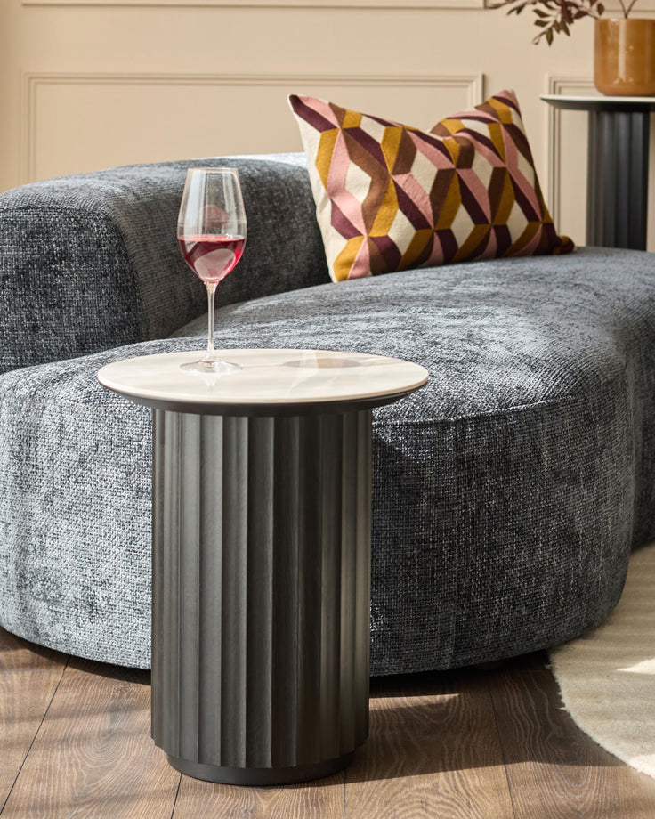 A modern lounge featuring a curved sofa, geometric cushion, side table with a glass of wine, and a round rug partially visible.