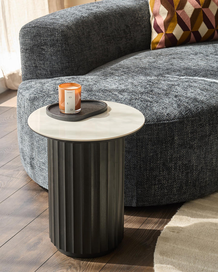 A stylish side table with a white top and black fluted base sits next to a modern grey sofa with a colourful geometric cushion.