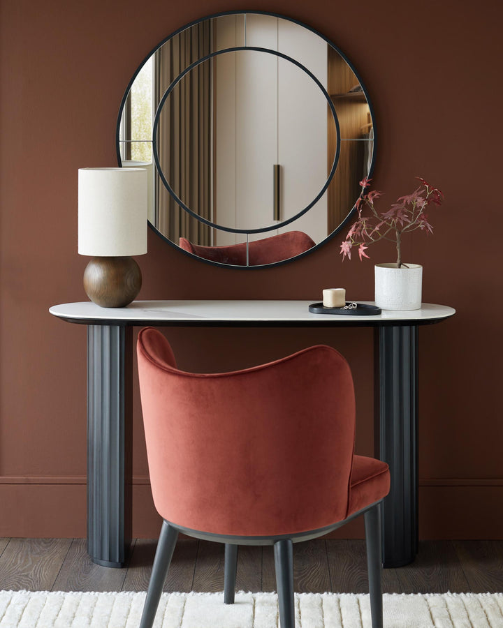 A dressing table with a round mirror, lamp, decor, and a velvet chair on a cream rug, creating an elegant and inviting space.