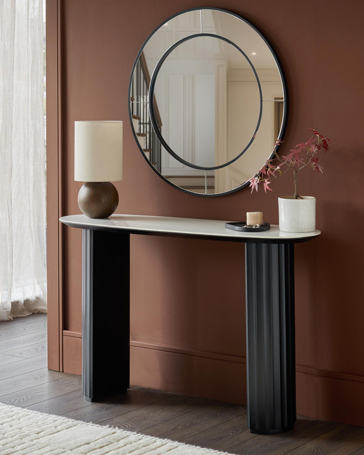 A console table with a round mirror above it, a lamp and a small potted plant on top. Interior design with wooden flooring.