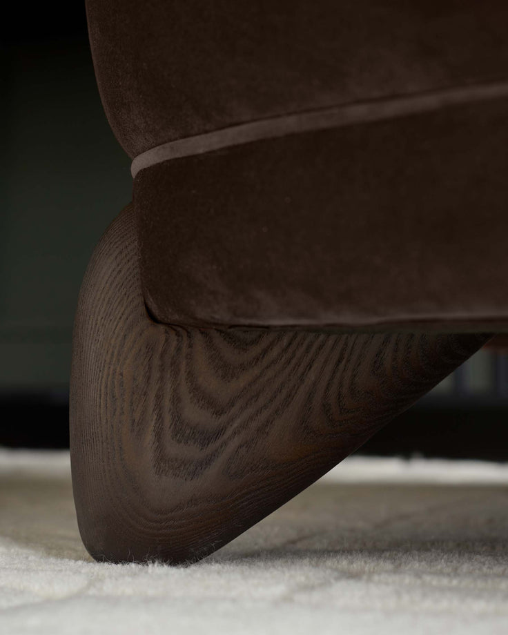Close-up of a brown velvet sofa with a dark wood leg, resting on a cream-coloured rug.