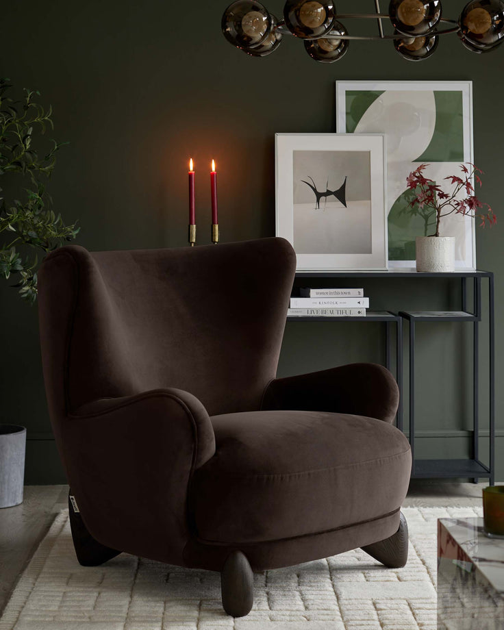A plush, brown velvet armchair sits on a cream rug. Behind it is a side table with books, artwork and candles.