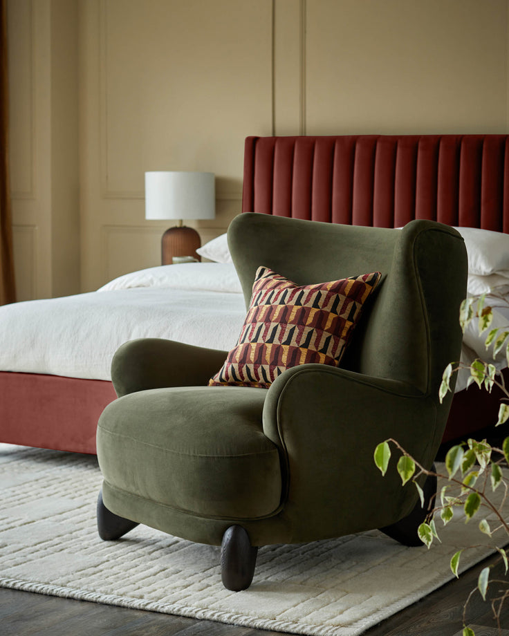 A stylish moss green armchair with patterned cushion sits on a rug beside a bed with a padded headboard.