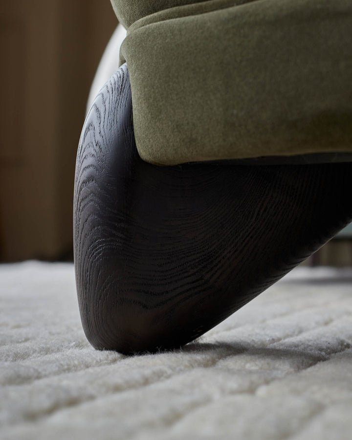 Close-up of the wooden leg and textured olive green fabric of a stylish sofa on a plush cream rug.
