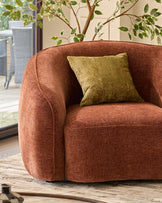 Harlene cushion in medium olive green, displayed on a rust-colored curved armchair in a bright interior space.