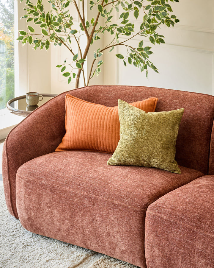 Harlene cushion in medium olive green displayed on a rust-colored curved armchair with orange pillow.