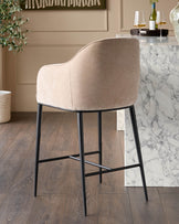 Champagne fabric bar stool with curved back, black metal legs and footrest, shown from rear angle at marble counter.