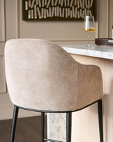 Champagne fabric bar stool with curved back, black legs, shown from rear angle at marble counter.