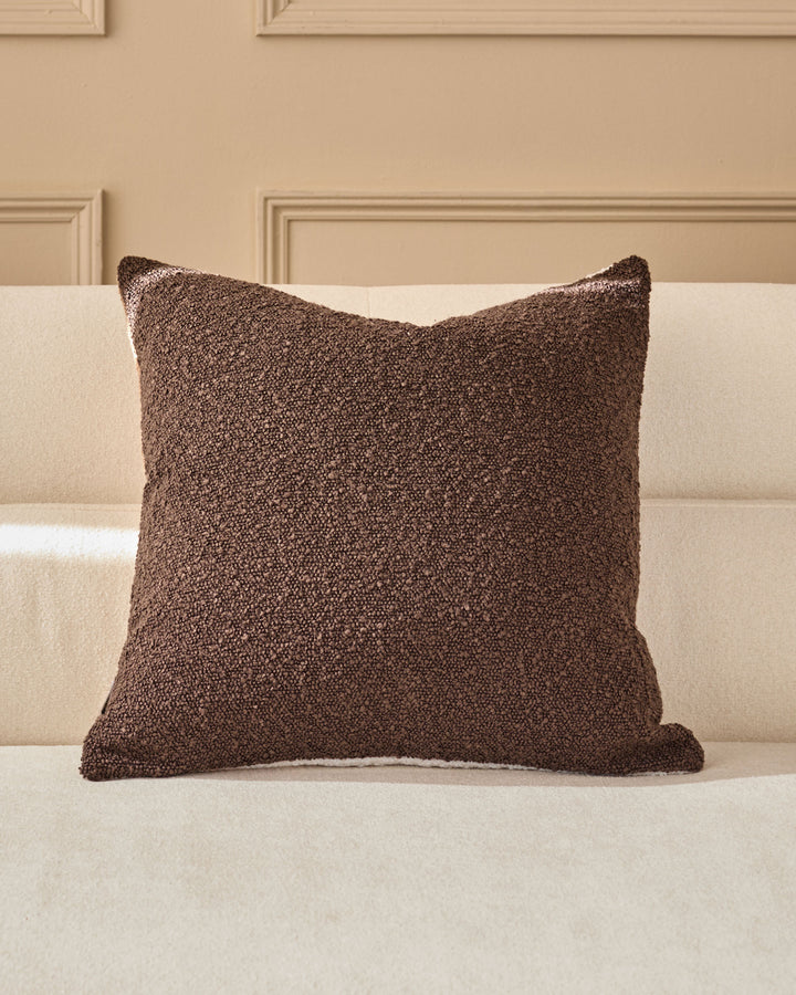 Brown textured cushion with piping on a cream upholstered sofa against a beige paneled wall.