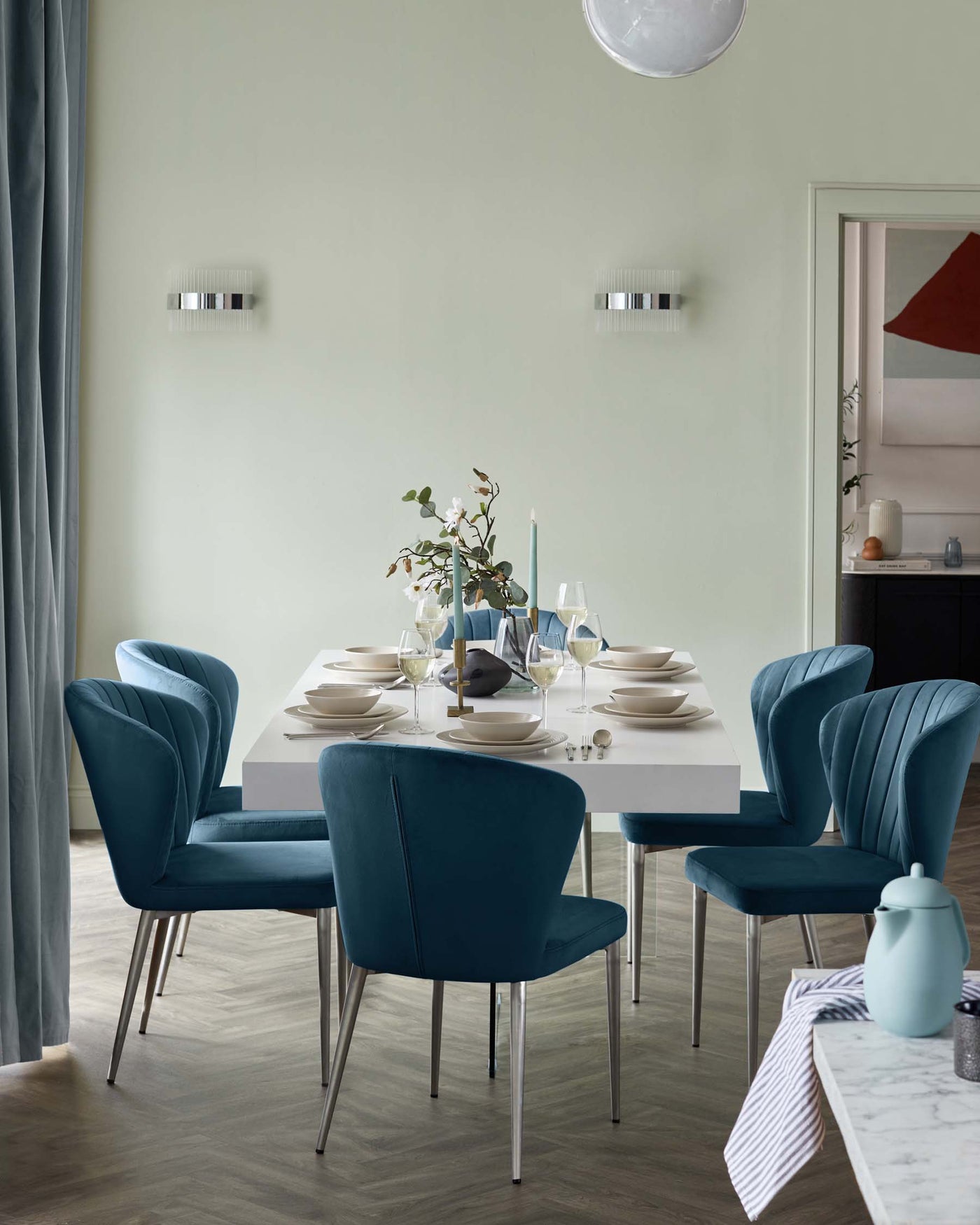 A contemporary dining room featuring an elegant white marble-topped table with a slender rectangular silhouette, paired with six luxurious teal blue upholstered chairs displaying curved backrests and vertical channel tufting. The chairs stand on sleek, slender metallic legs, complementing the table's clean lines. The set exudes a modern yet timeless appeal, perfect for both casual and formal gatherings.