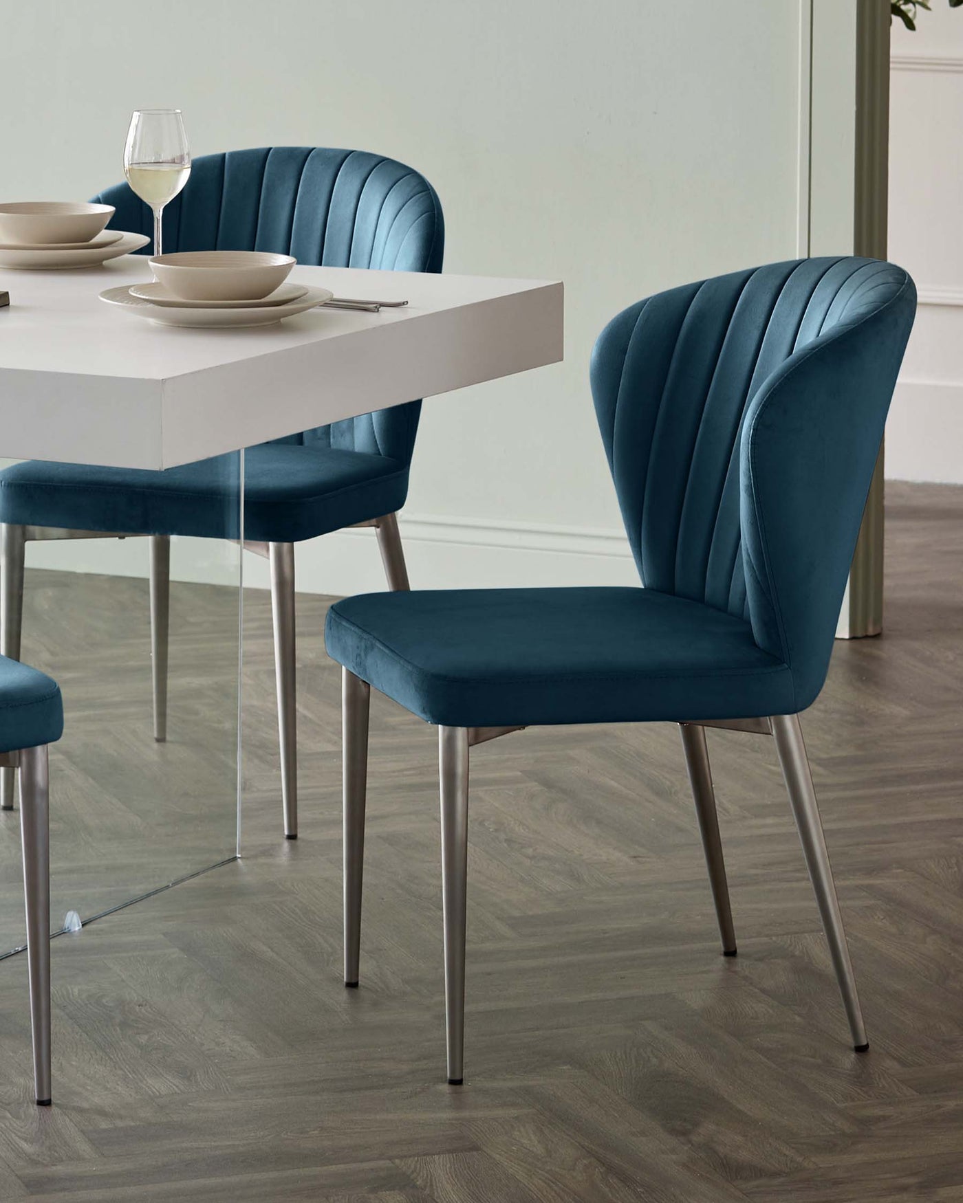 A contemporary dining room set featuring a white rectangular table with a simple, sleek design and two blue velvet upholstered chairs with a vertical channel-tufted backrest and cushioned seat, all supported by metallic silver legs.
