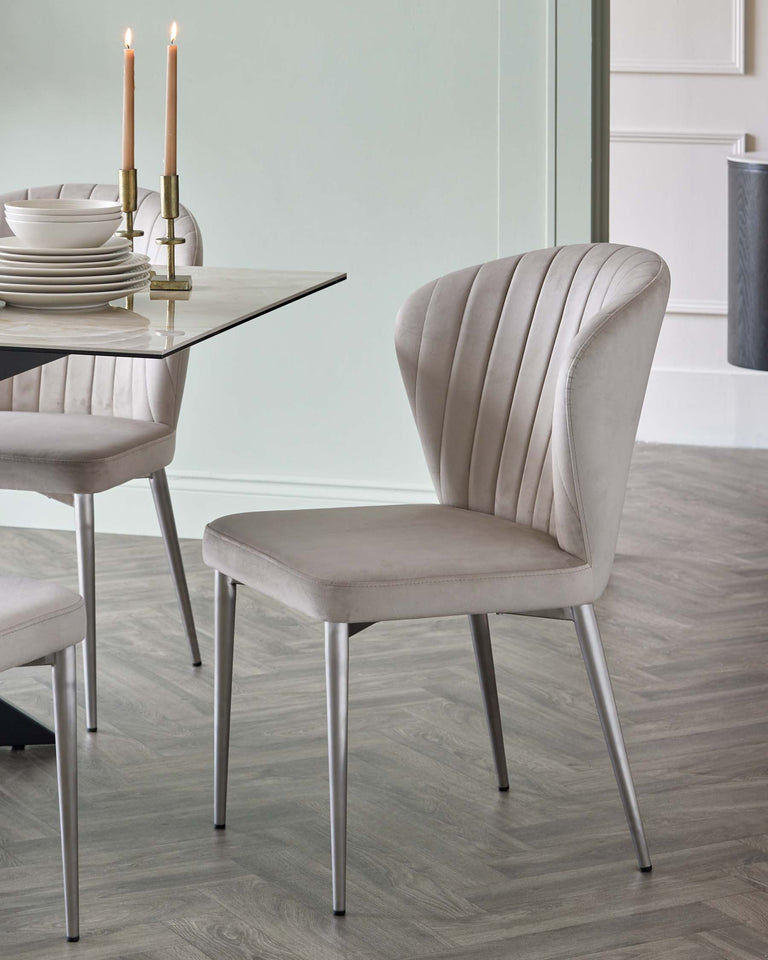 Elegant dining chair with a curved back design and plush vertical channel tufting in a soft cream upholstery. Features a smooth metal base with slim, tapered legs in a brushed finish. A portion of a contemporary glass-top dining table with a set of plates and candle holders is visible in the background.
