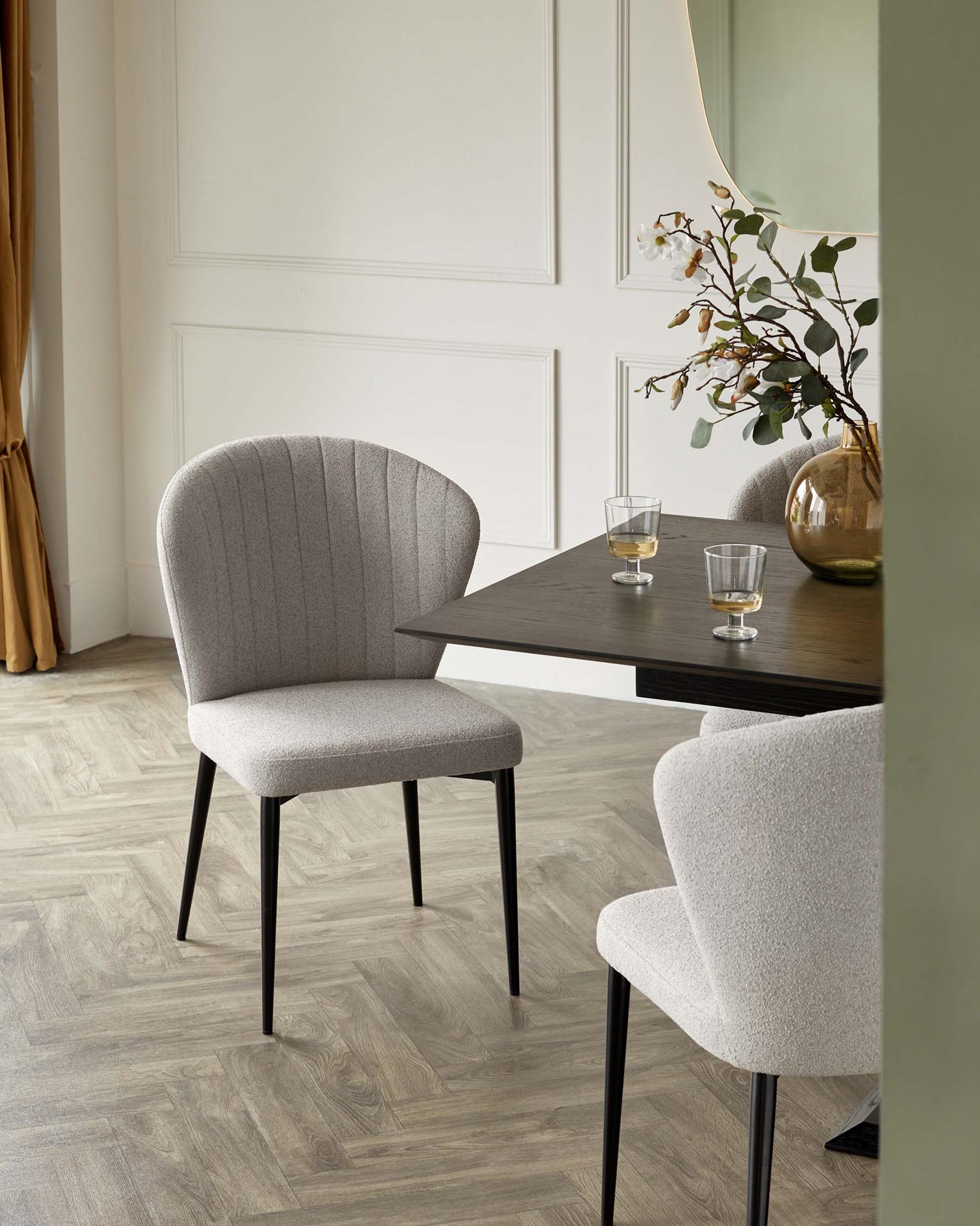 Modern minimalist dining setup featuring an elegant dark wood table paired with two stylish upholstered chairs with channel tufting and black legs. The design is complemented by a gold-rimmed mirror and vase with greenery.