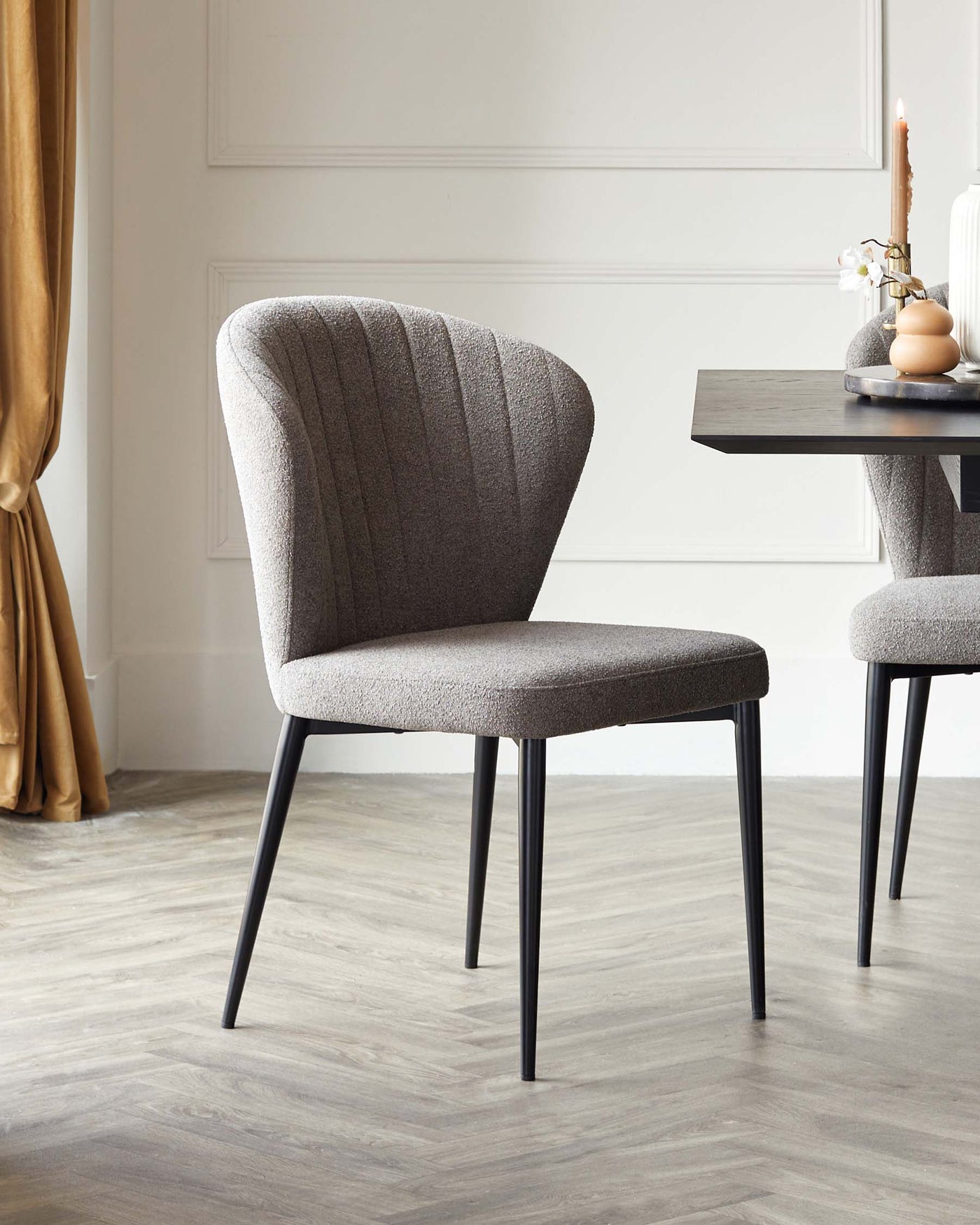 Modern upholstered dining chair with vertical stitching detail on the backrest, a textured grey fabric, and angled black metal legs. A minimalist rectangular black dining table is partially visible with decorative items on top. The setting features a light hardwood floor and elegant white wainscoting on the walls, accented by a golden-coloured curtain on the left side.