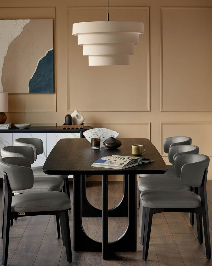 A modern dining set featuring a dark wood table and six grey leather chairs, illuminated by a tiered cream pendant light.