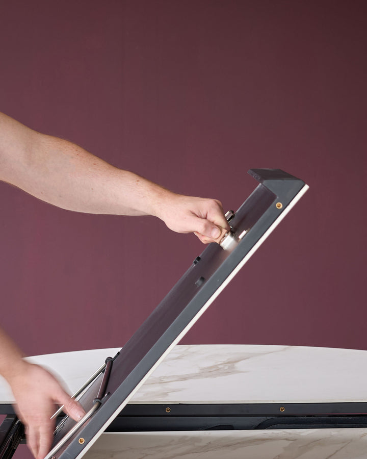 Hand adjusting extension mechanism on natural ceramic dining table with dark frame against burgundy background.