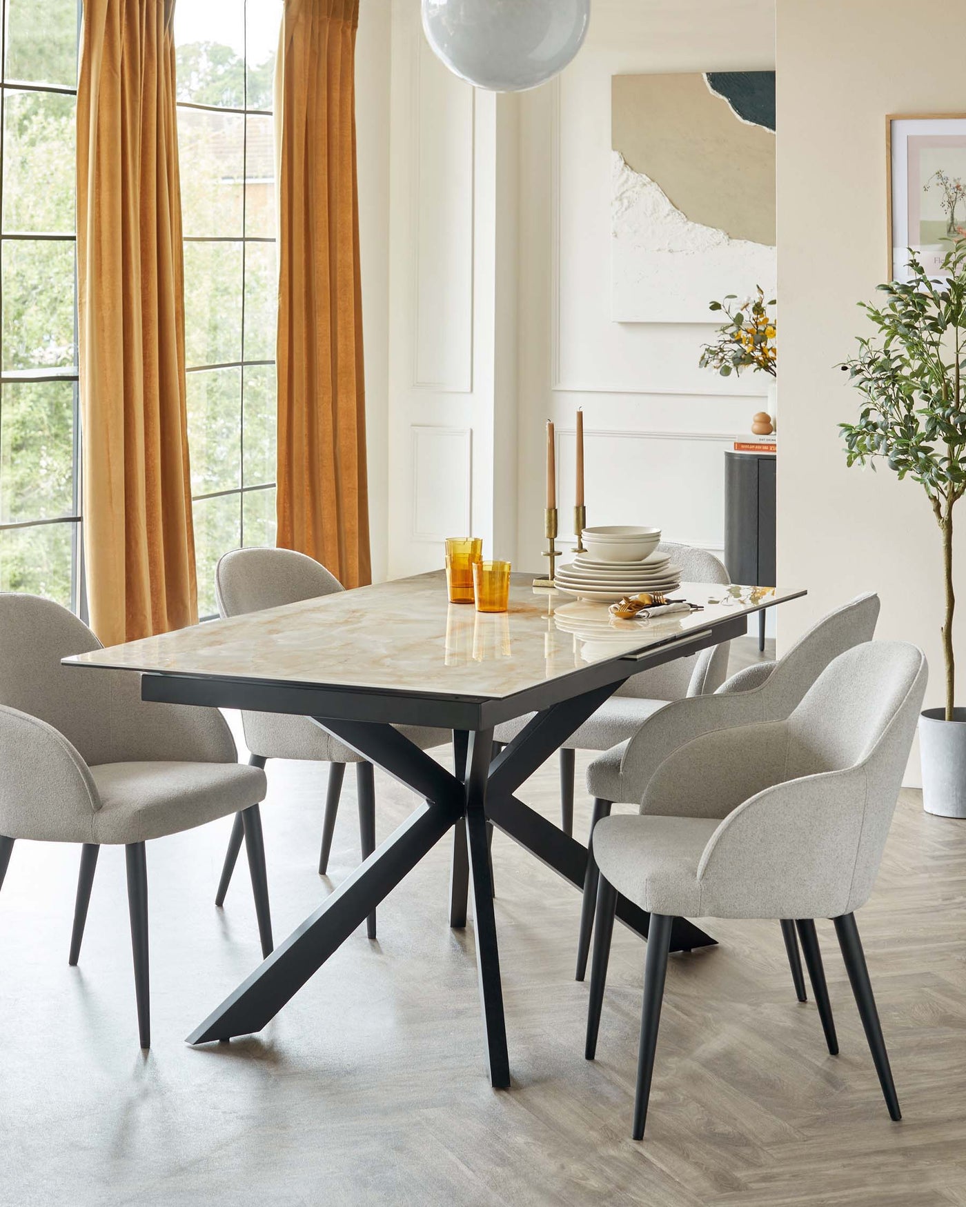 Modern dining room with a rectangular marble-top table featuring a geometric black metal base. Accompanied by four elegant, light grey upholstered chairs with a smooth, curved design and black legs. Warm natural light filters through large windows with golden curtains accentuating the space.
