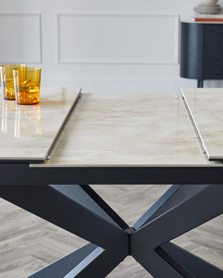 Modern dining table with a polished marble top featuring creamy hues and a sleek surface. The base is an intersecting matte black metal frame with a dynamic, geometric design, providing sturdy support with a contemporary edge. Two decorative amber-coloured glasses rest on top.