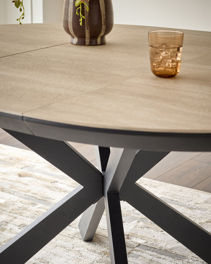 Hayden round extending dining table in natural travertine with modern crossed-leg base, styled with amber glass and potted plant.