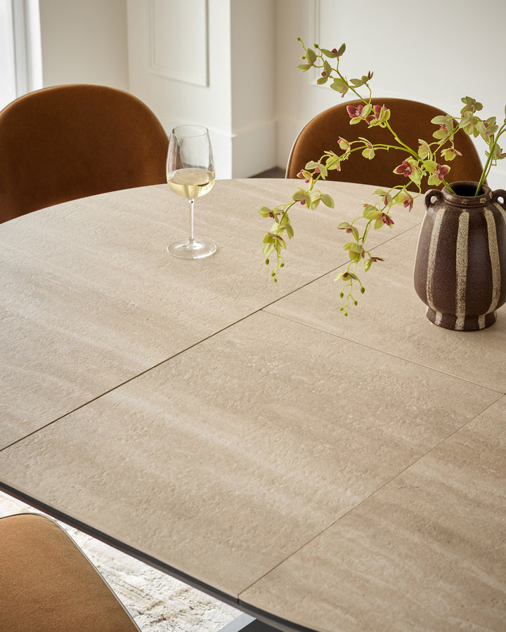 Hayden round extending dining table in natural travertine ceramic, shown extended with place setting and floral centerpiece.