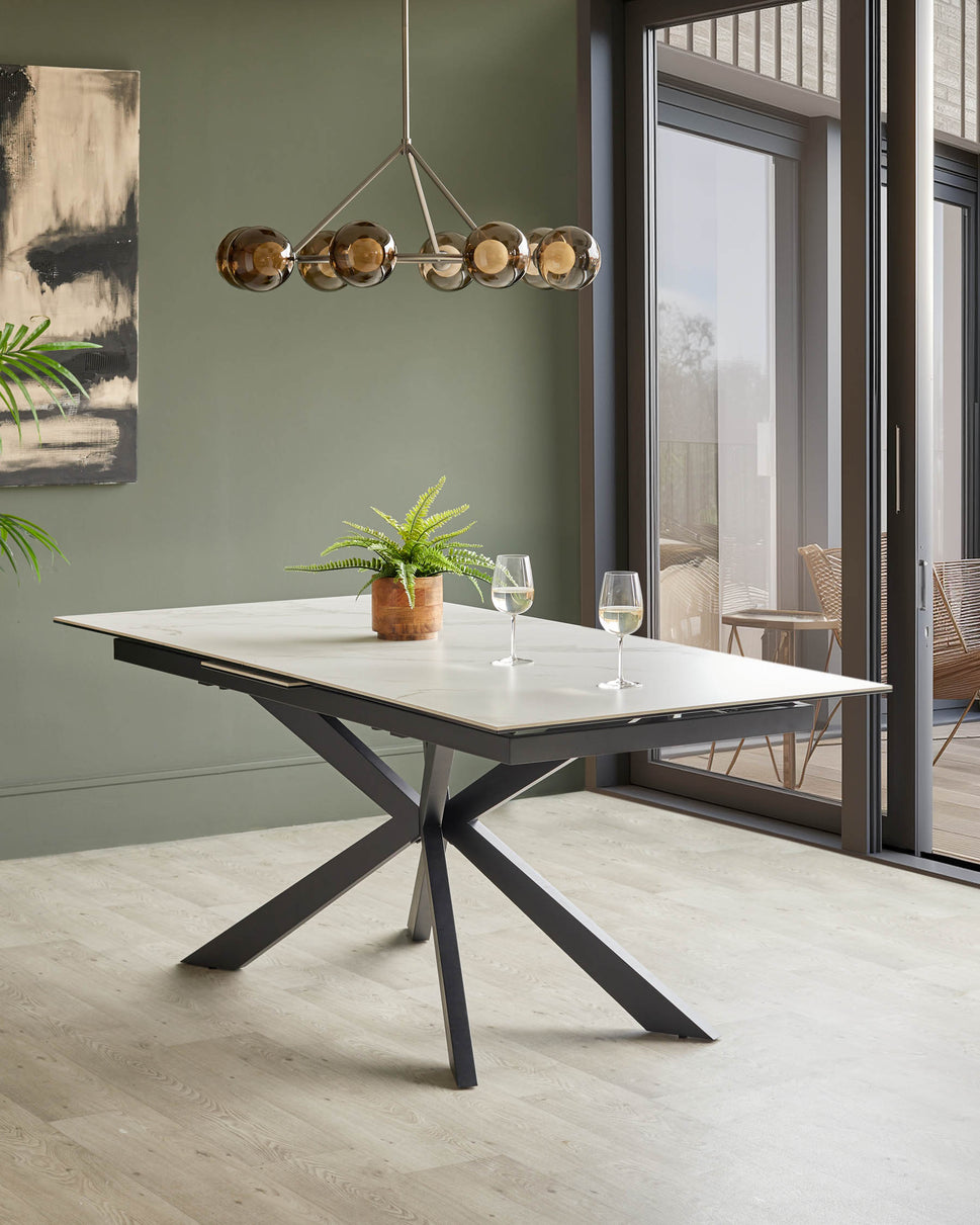 A modern dining table with a white top and dark metal base, set with wine glasses and a fern.