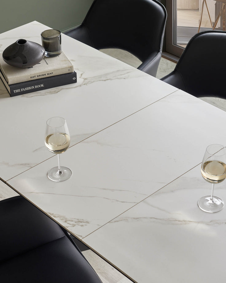 A modern marble dining table, styled with wine glasses, books, and a black ceramic vase and candle. Features black leather chairs.