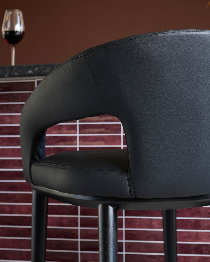 Close-up of a black leather bar stool, featuring a curved back and sleek design. A glass of red wine is blurred in the background.
