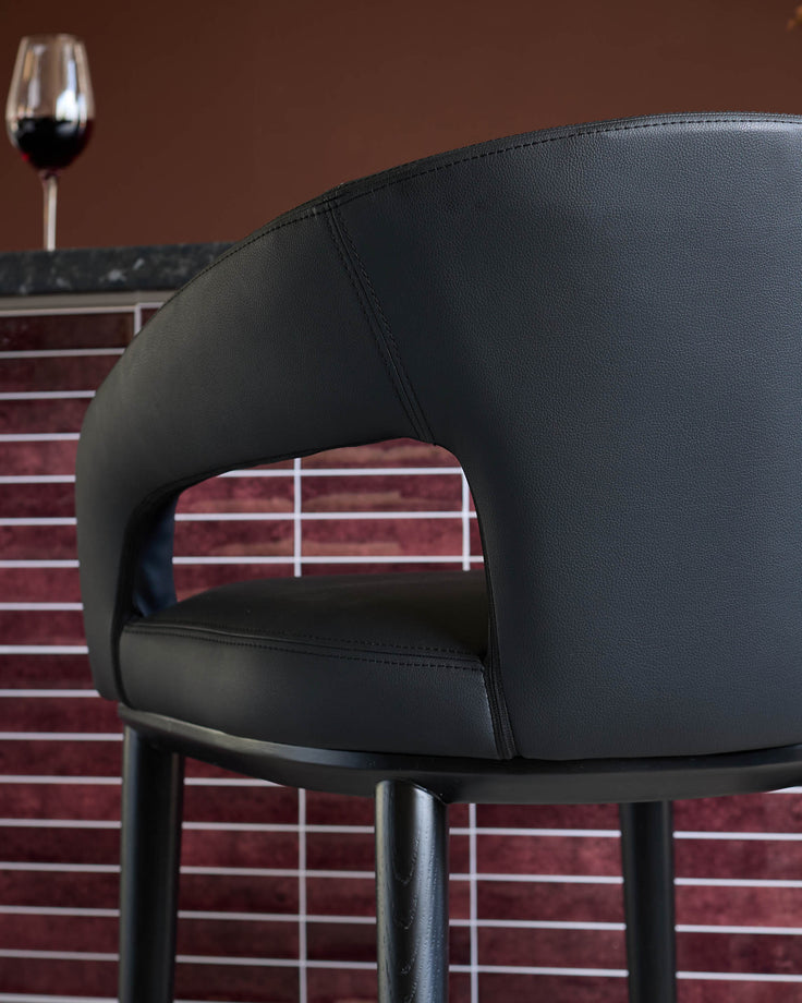 Close-up of a black leather bar stool, featuring a curved back and sleek design. A glass of red wine is blurred in the background.